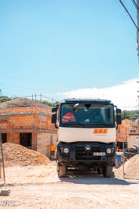 Ouvrier en bâtiment posant des briques rouges sur un mur en construction, portant casque de chantier jaune, lunettes de protection et t-shirt rouge